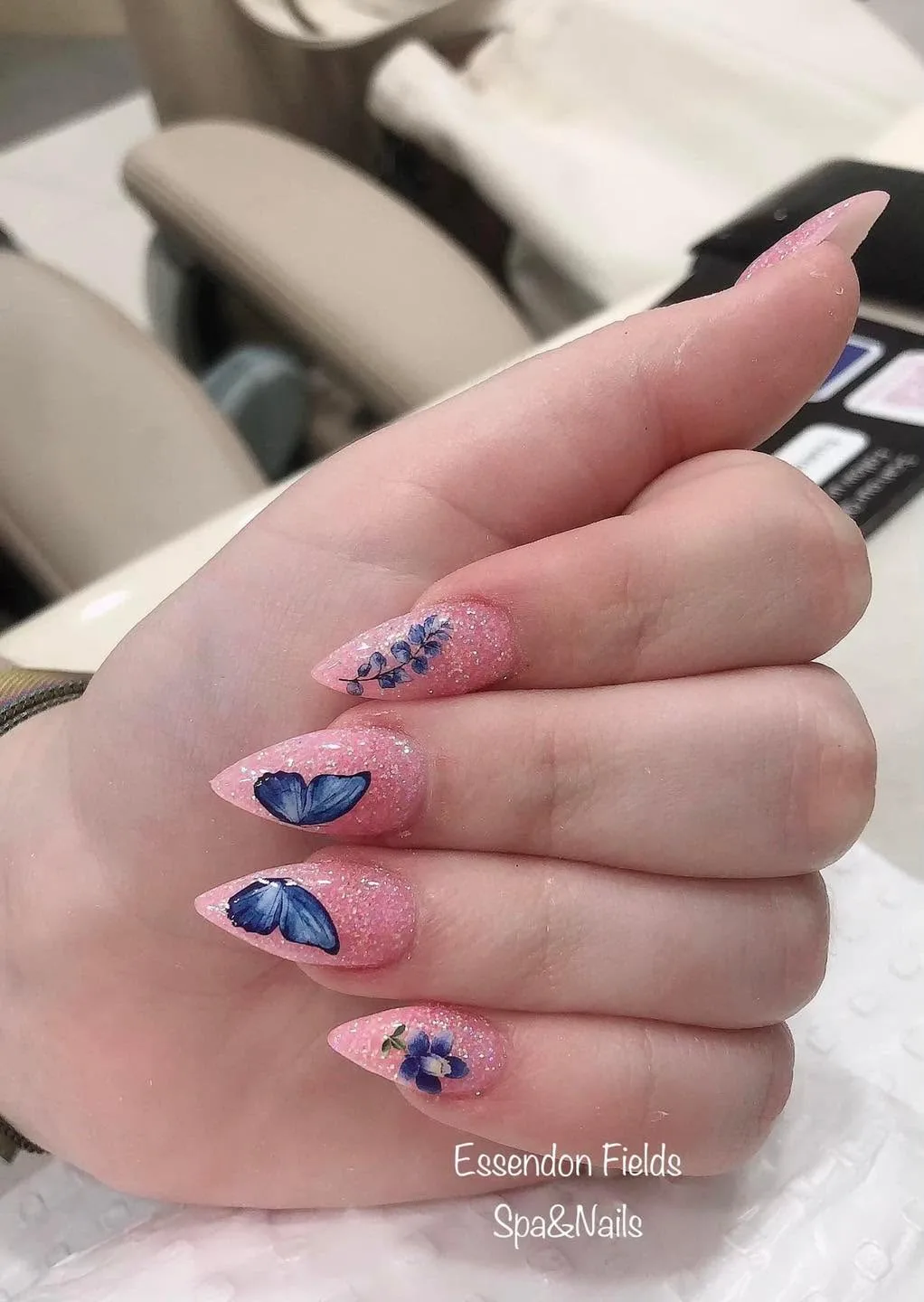 Stiletto nails in pink glitter with blue butterfly-wing and blue floral art, on a cream chair
