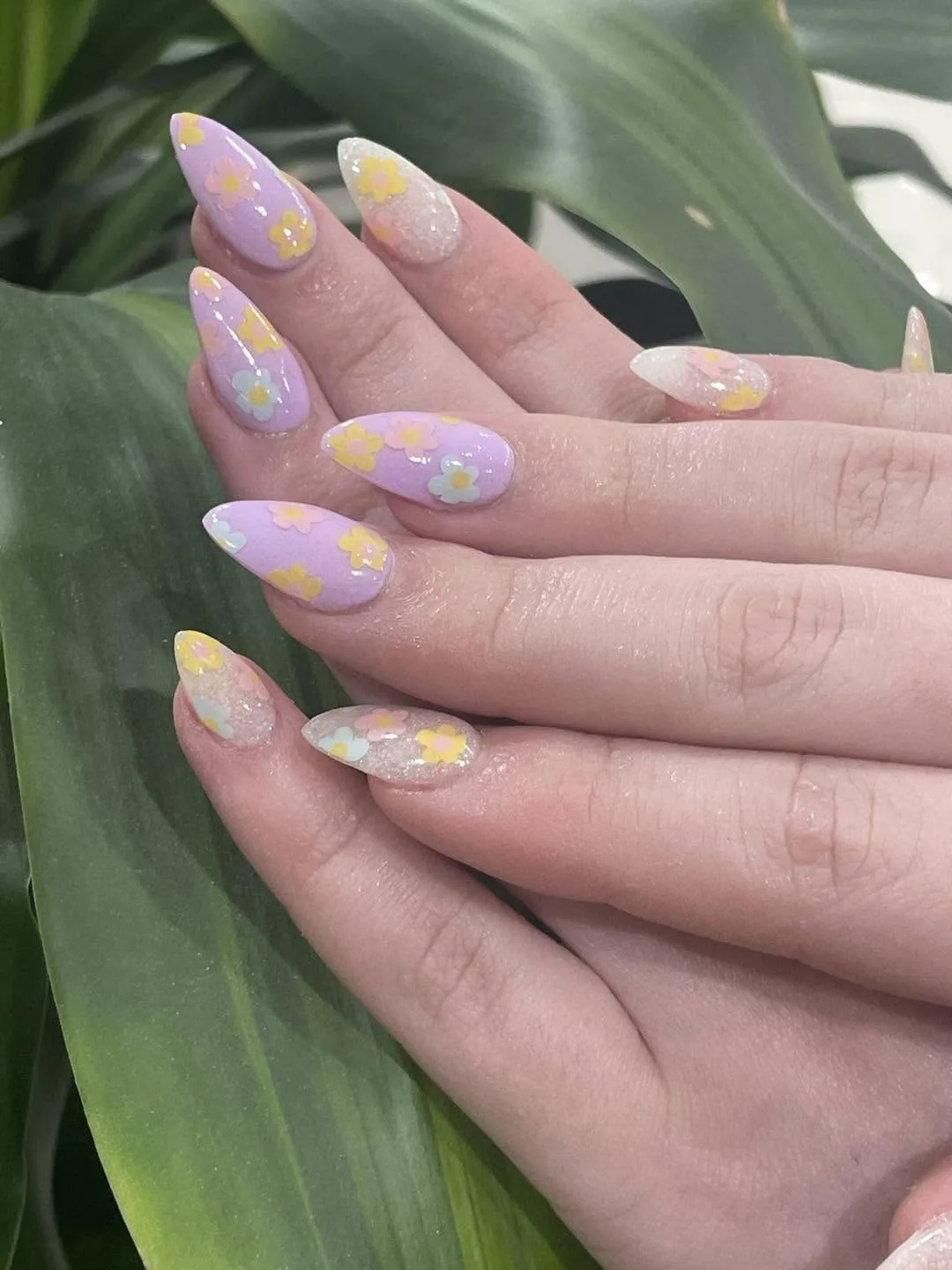 Almond nails in lilac with retro yellow and white daisy art and clear glitter accents, against a green leaf
