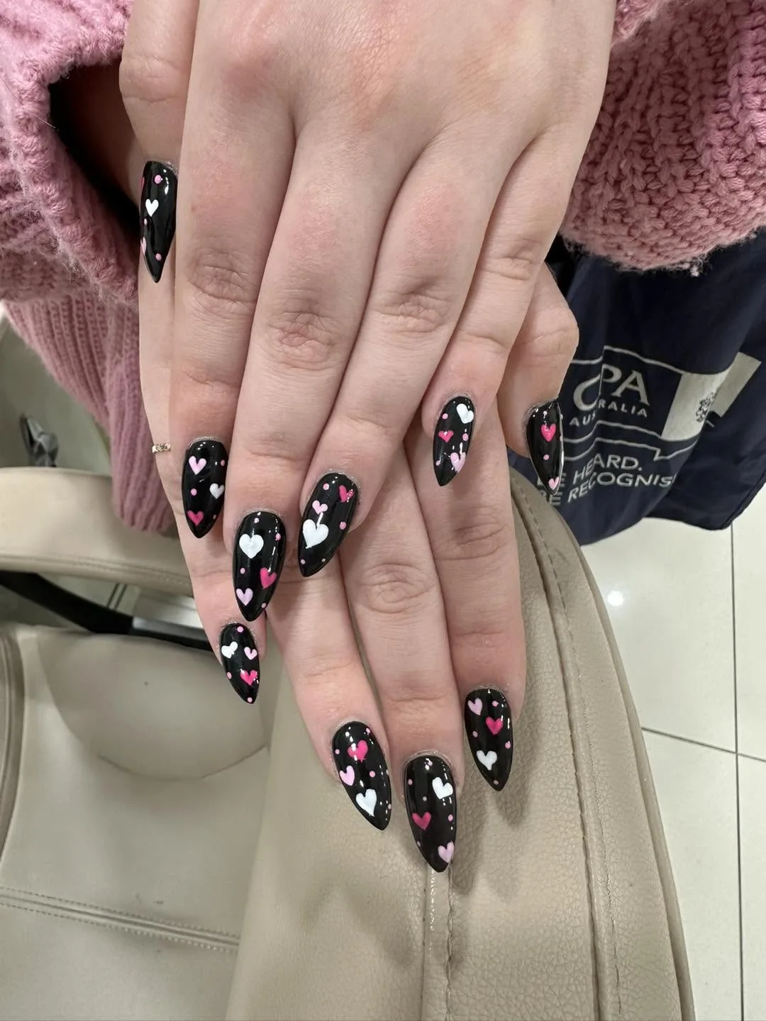Almond black nails with pink and white hand-painted heart art, glossy, resting on a cream salon chair