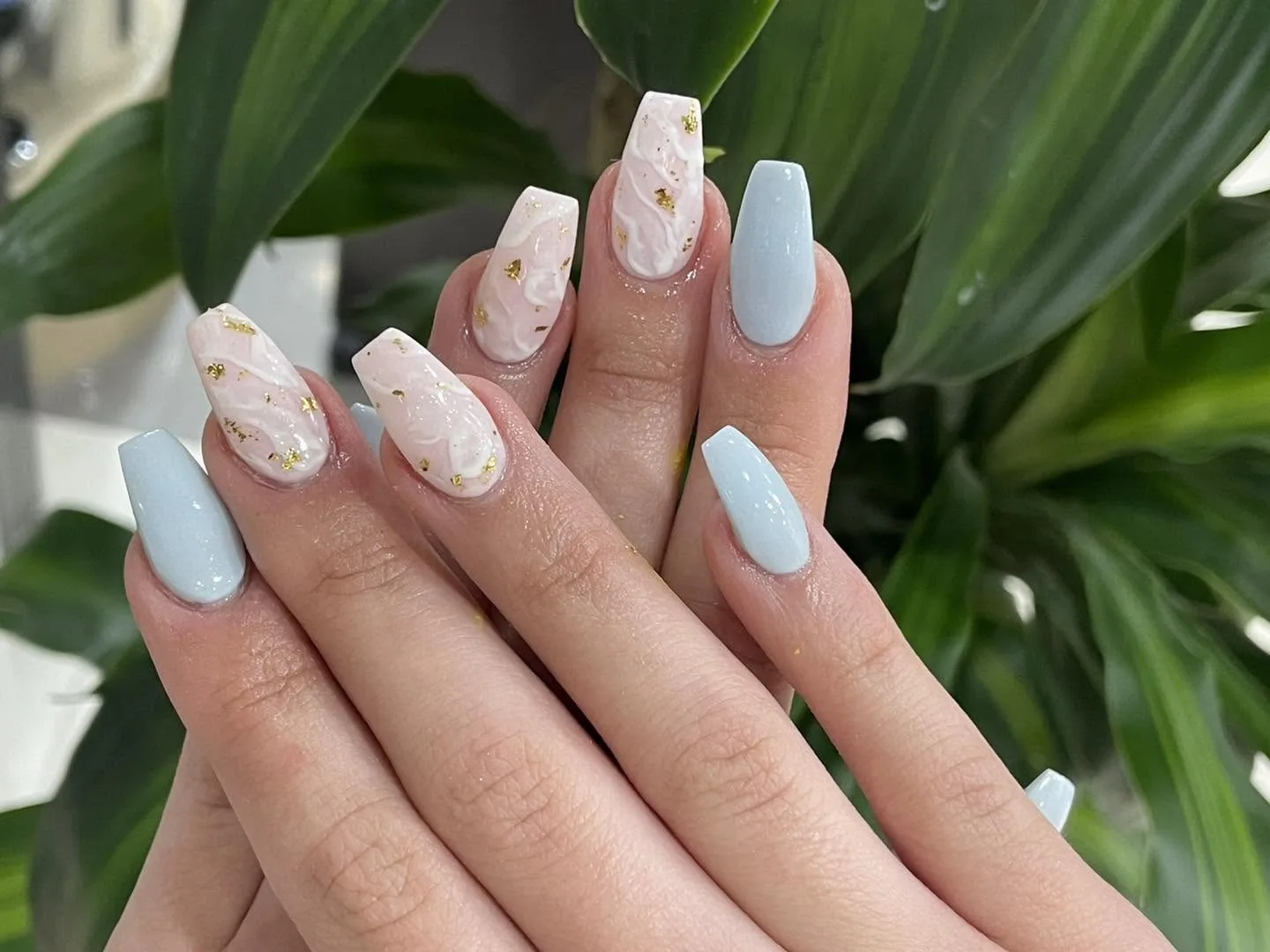 Coffin nails in baby-blue and blush-marble with gold-flake accents, glossy, cradled against lush dracaena foliage