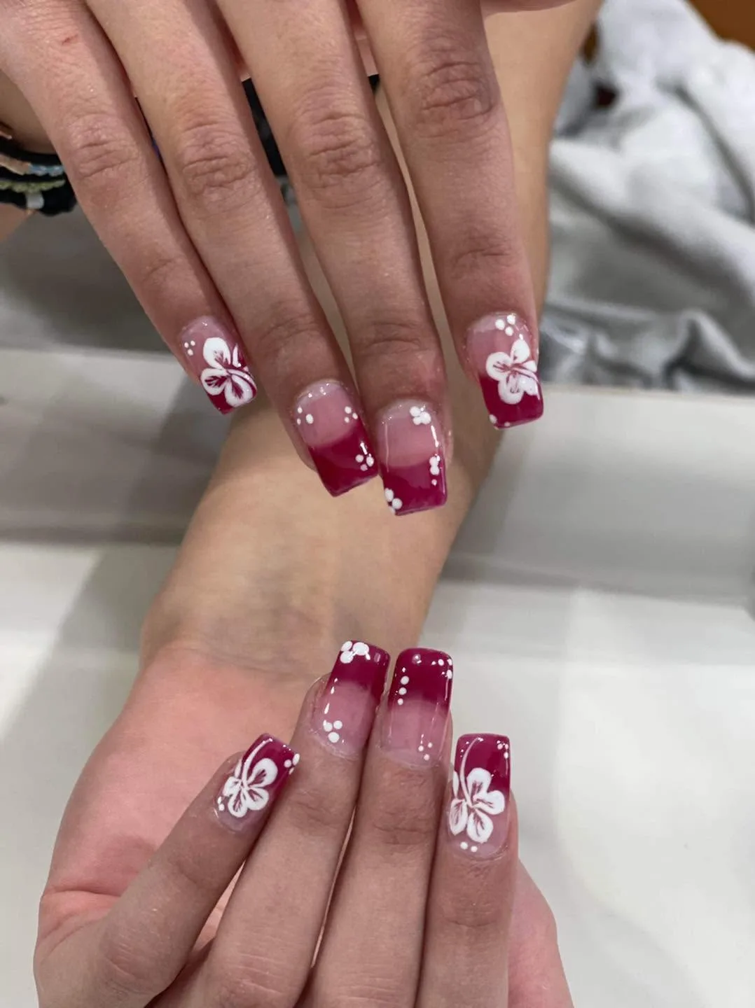Two hands with square deep-magenta nails featuring white hibiscus-flower art and dot accents, on a white surface