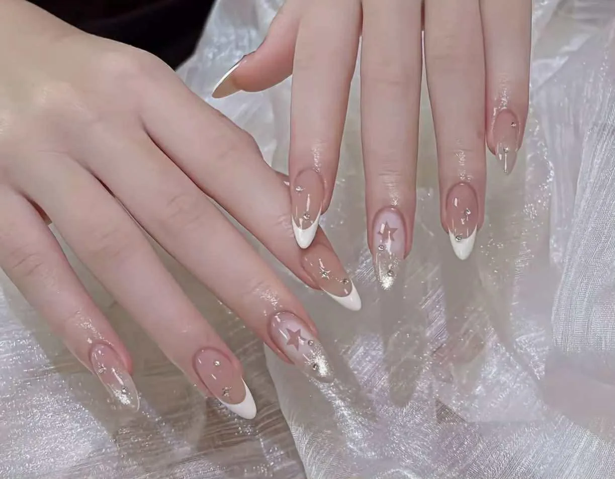 Almond nude nails with white French V-tips, star art, rhinestones and gold-foil accents, on iridescent cellophane