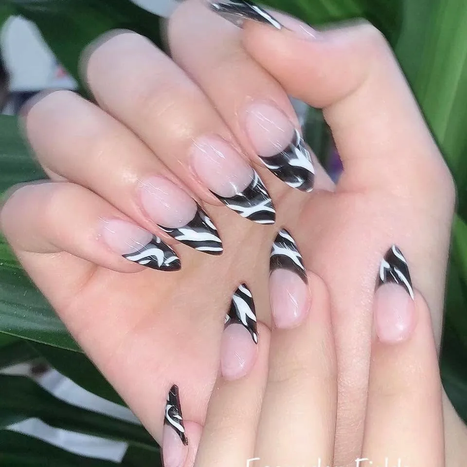 Long stiletto nails in sheer nude with black-and-white zebra-swirl French tips, glossy, on green palm-leaf backdrop
